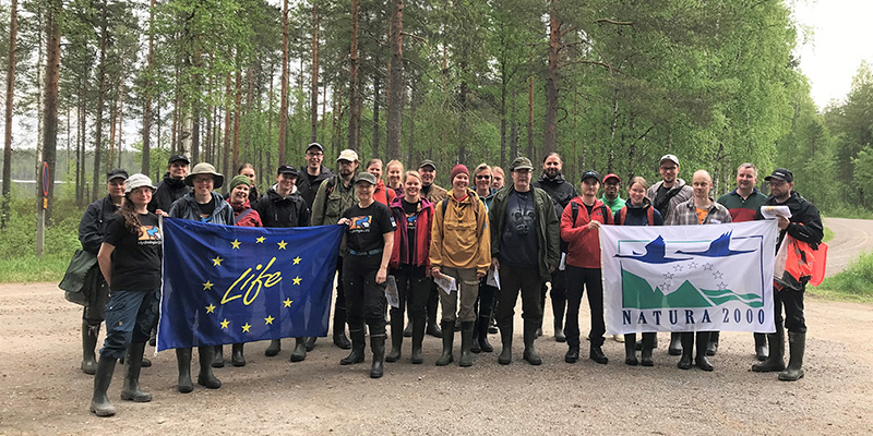 Ryhmä ihmisiä seisoo ulkona rinnakkain ja katsoo kameraan, etummaiset kannattelevat suuria Life- ja Natura 2000 -lippuja.