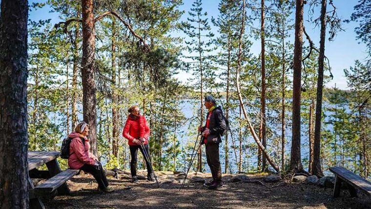 Kolme retkeilijää taukopaikalla puiden katveessa. Yksi istuu penkillä. Taustalla on järvi.