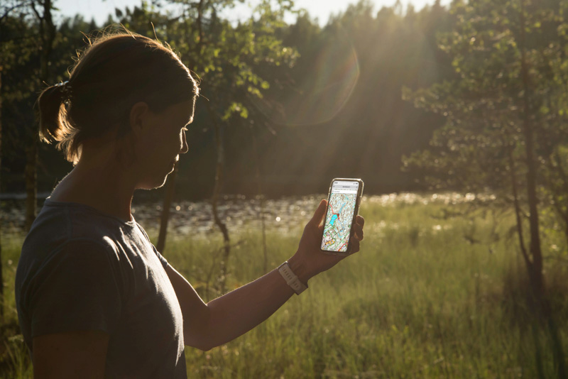 En kvinna står i naturen och tittar på mobilen i handen.