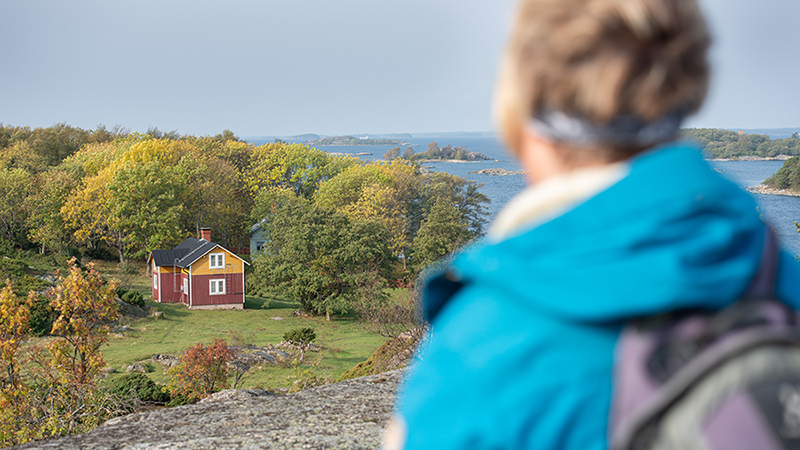 En person står på ett berg och tittar nedåt till stranden där det finns ett gammalt trähus med trädplan och lövträd. I bakgrunden havet.