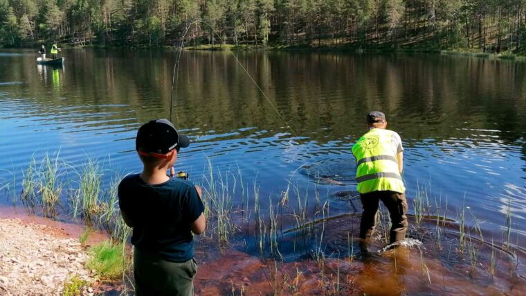 Poika seisoo rannalla virveli k&auml;dess&auml;&auml;n, siima on kire&auml;ll&auml;. Mies keltaisessa liiviss&auml; on kahlannut veteen.