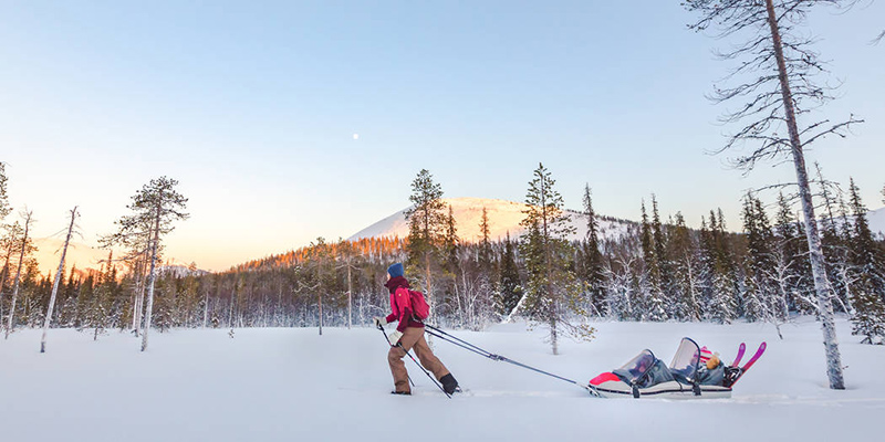 Talvinen tunturimaisema. Aurinko valaisee taustalla tunturihuiput. Retkeilijä hiihtää umpihangessa vetäen perässään ahkiota, jossa on kaksi lasta.