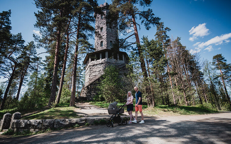 Nainen ja mies kävelevät rinnakkain korkean kivitornin edessä polulla aurinkoisena kevätpäivänä, nainen työntää lastenvaunuja.