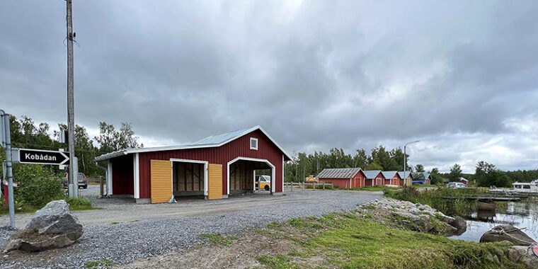 Harjakattoinen puurakennus meren rannalla soratien vieressä, rakennuksen etusivu katoksen muotoinen, kaksi isoa sisäänkäyntiaukkoa.