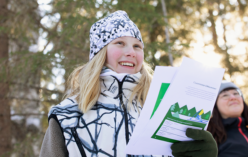 Hymyilevä poika ulkona metsässä kädessään tehtävälappuja. Taustalla hymyilevä nainen.