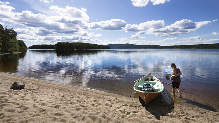 En roddbåt och människa bredvid på en sandstrand i en solig sommardag. På bakgrunden en sjö och stränder med skog.