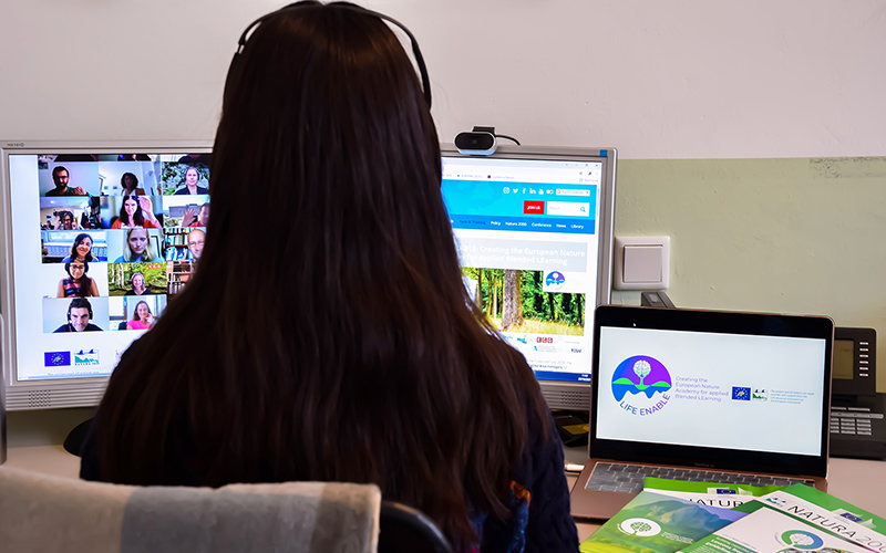 A longhaired person, back turned to camera, sits at a desk looking at three computer monitors.