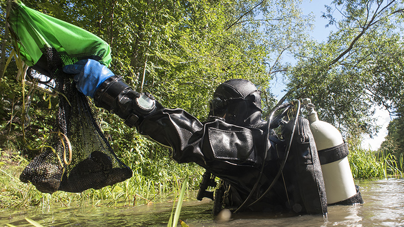 A person in a diving suit stands in a river and hands out a bag made of a net with mussles in it.