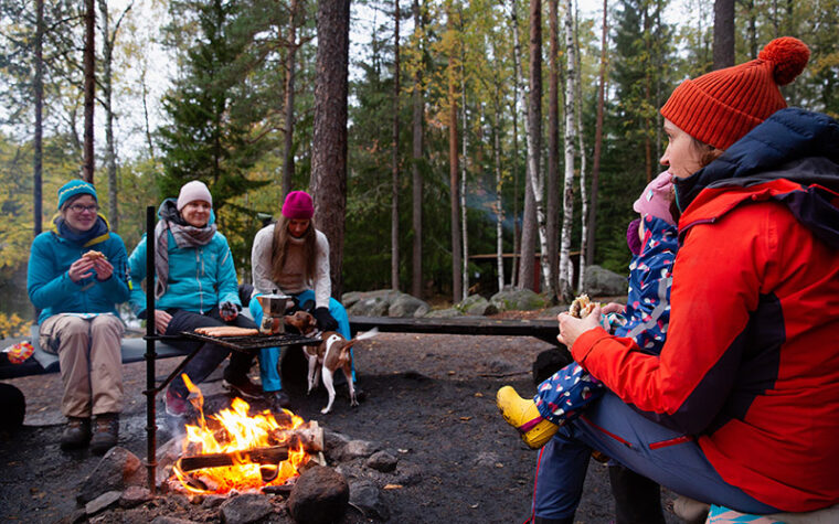 Ihmisiä istumassa takit päällä ja pipot päässä tulipaikan ympärillä. Nuotiolla paistumassa makkaroita.