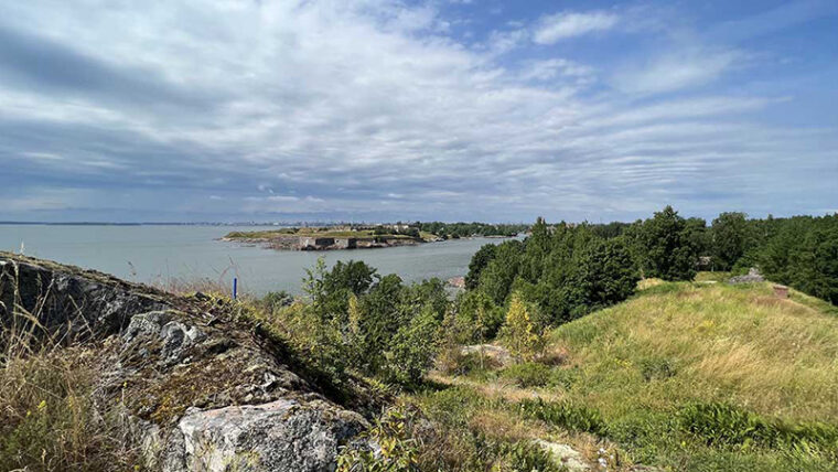 Kalliota, ruohopeitteisiä kumpuja ja lehtipuita meren rannalla, salmen takana vastarannalla kivisiä linnoitusmuureja.