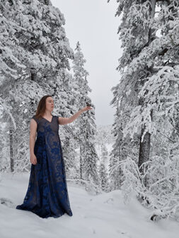 Nainen seisoo lumisessa metsässä pitkähelmaisessa, hihattomassa juhlamekossa ja ojentaa kättään, käden päällä pieni lintu.