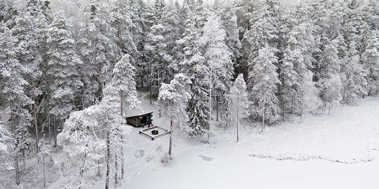 Luminen maisema ilmasta kuvattuna, rannalla puiden keskellä hirsikota ja sen edessä nuotiopaikka puupenkkien ympäröimänä.