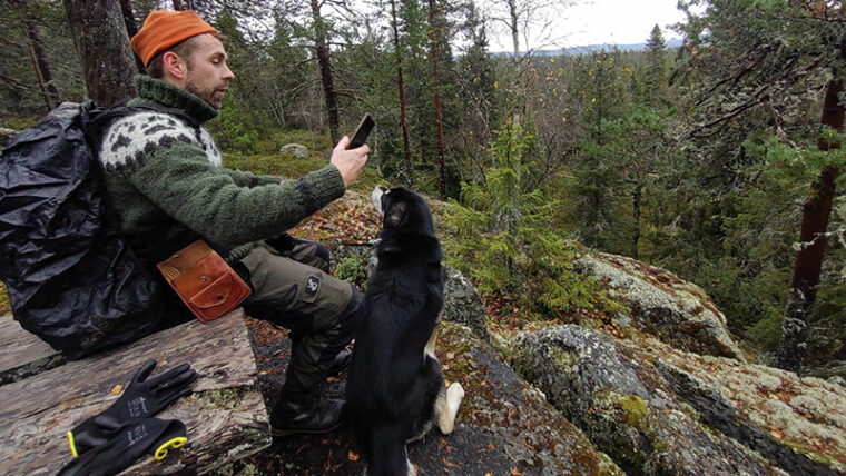 Mies istuu kallion reunalla metsässä ja katsoo puhelintaan, koira istuu vieressä.