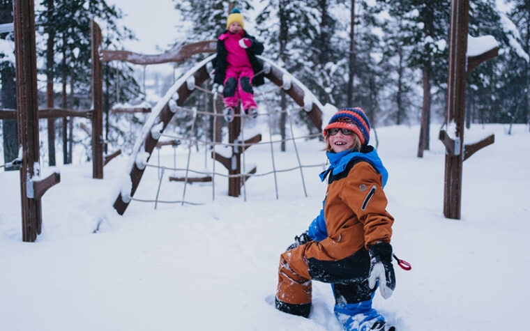 Kaksi lasta leikkii lumihangessa metsämaisemassa, toinen on kiivennyt puusta ja verkosta tehdyn lasten kiipeilytelineen päälle.