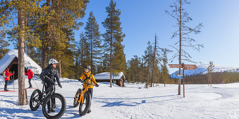 Aurinkoinen talvipäivä, kaksi hiihtäjää ja kaksi maastopyöräilijää seisoo kotarakennusten luona, taustalla tunturi.