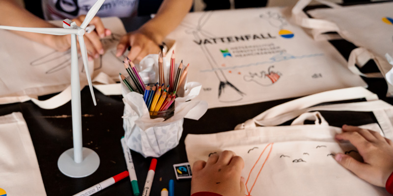 There are cloth bags and markers on the table. The picture also shows hands that are drawing on the bags.