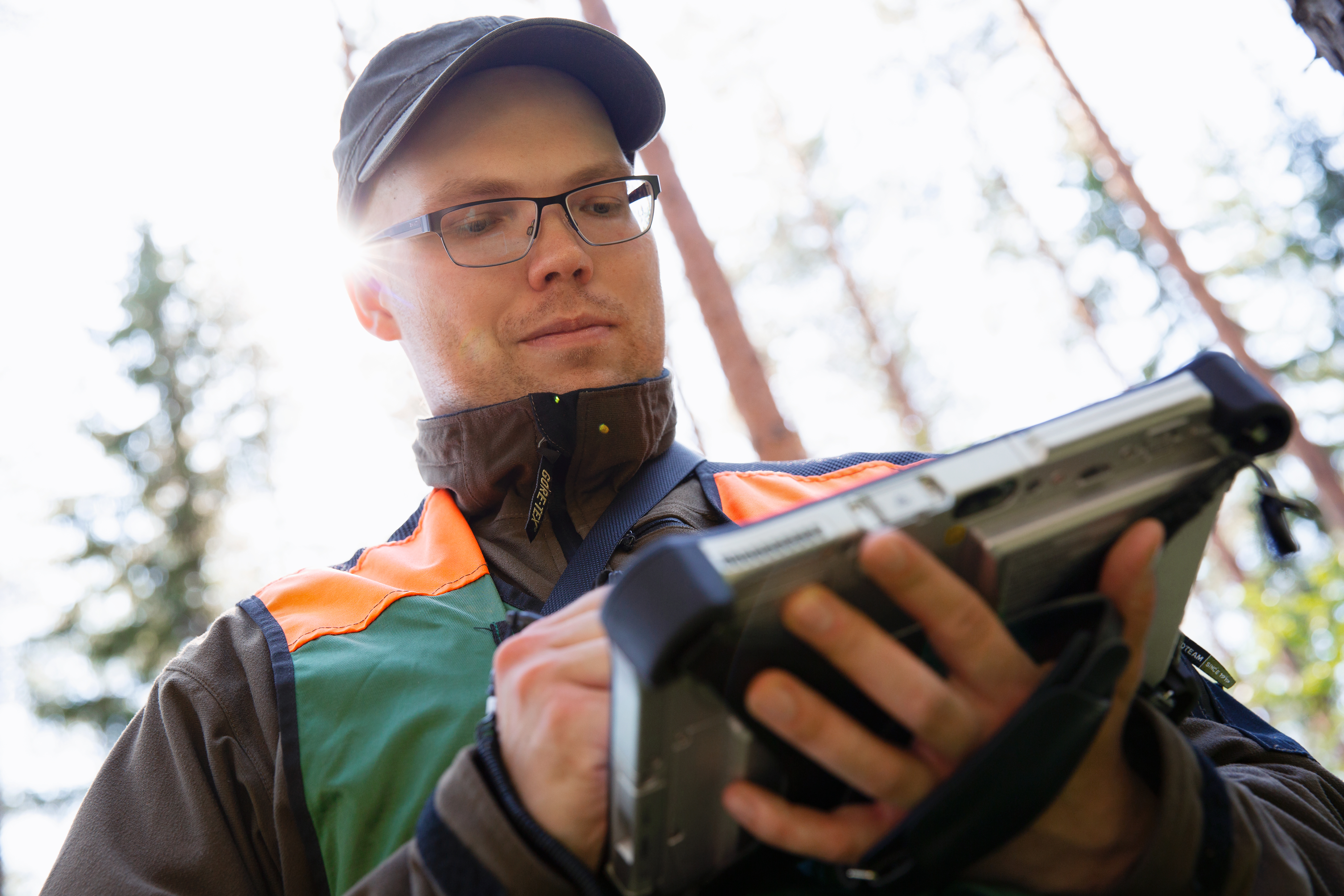 Metsäasiantuntija kesäiessä metsässä alaviistosta kuvattuna niin että hänellä on toisessa kädessä maastotallennin johon hän tekee muistiinpanoja.