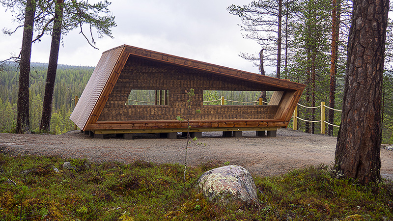 A wooden, open shelter with windows on a hilltop facing a forest view.