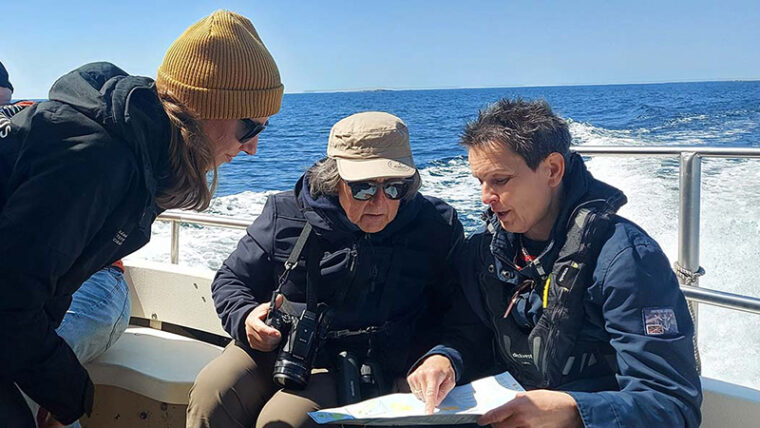 Tre kvinnor i en båt på havet, en av dem visar på kartan till en annan, som har en kamera.