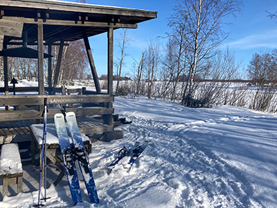 Solig vinterdag vid havstrand med snö. Korta skidor lutar mot en träskydd med bord och bänkar