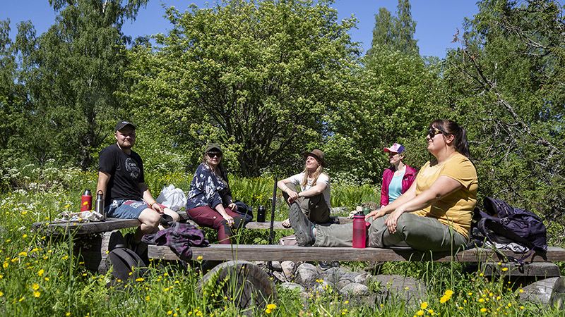 Människor sitter på träbänkar på en äng på en solig sommardag, bakom dem lövträd.