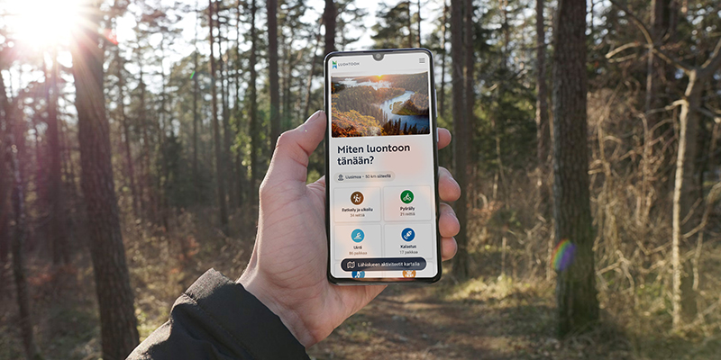 En hand håller i en smarttelefon på vars skärm man ser texten ”Miten luontoon tänään?”, en bild av ett ålandskap samt ikoner för olika naturhobbyer. I bakgrunden syns träd som solen skiner mellan.