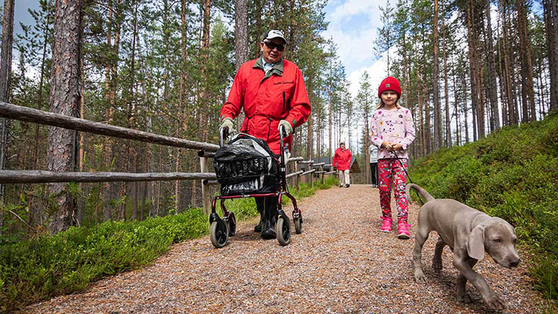 Mies kävelee rollaattorin kanssa tasaisella metsäpolulla. Hänen vierellään kävelee tyttö, joka taluttaa koiraa.