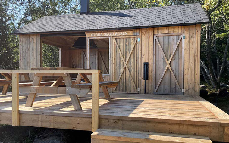En träbyggnad på en terrass av brädor med en bordsbänk. Bakom terrassen en kokskjul och två rum med dörrar.