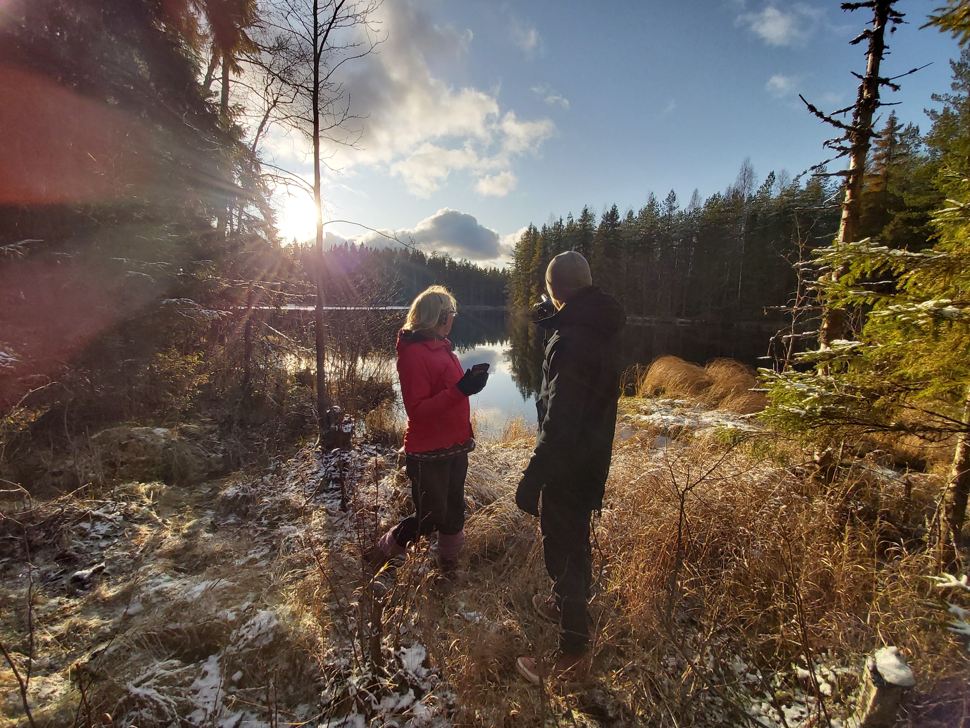 Maisemakuva, jossa kaksi henkilöä seisoo järven rannalla ja katsovat järven suuntaan, jossa paistaa aurinko.