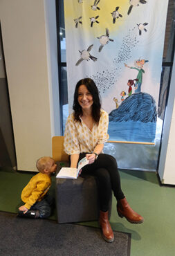 A smmiling woman sits on a chair with an opened bok. A child kneeling on the floor beside the chair looks at her and smiles.