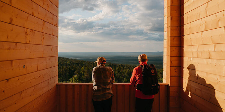 Kaksi ihmistä nojaa puurakennuksen näköalapaikan kaiteeseen ja katselee metsä- ja tunturimaisemaa.