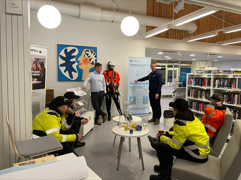 Fyra ungdomar klädda i orange och gula arbetskläder tittar och lyssnar när två män presenterar vindkraftverkens teknik i biblioteket.