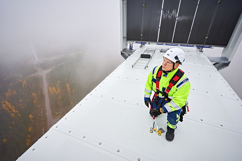 A man dressed in protective clothing and a safety harness on top of the engine room of the wind turbine. Below you can see a forest in brown colors.