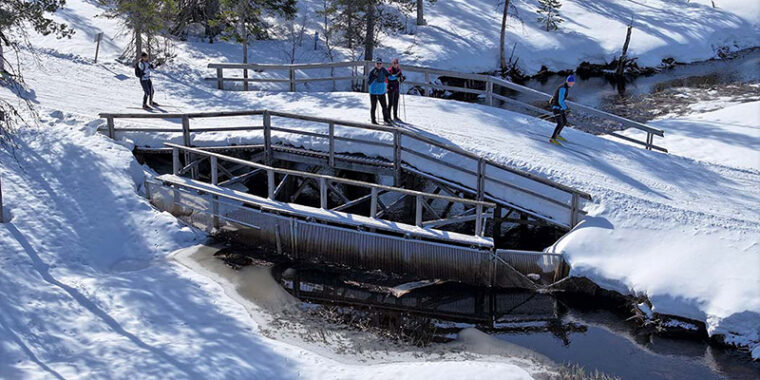 Lumen peittämä matala kaarisilta, jonka alla sula joki. Sillan päällä latuja ja hiihtäjiä, sillan alla puinen seinämä joen poikki.