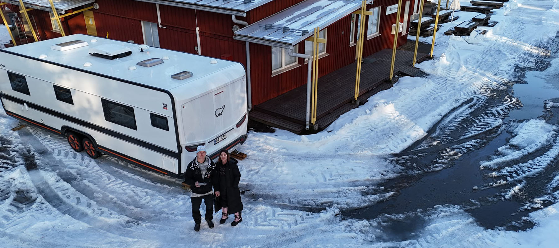 Mies ja nainen asuntovaunun kanssa leirintäalueen päärakennuksen vieressä. Piha on vielä luminen,