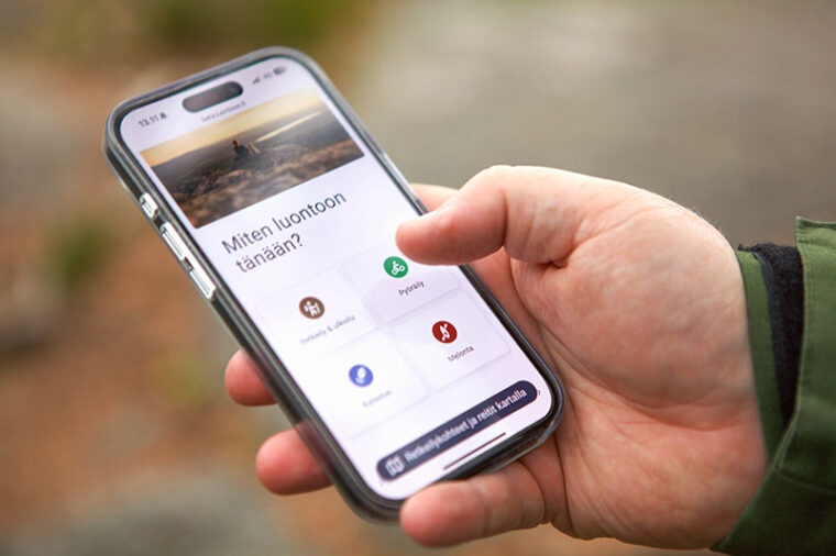 Hand holding a smartphone with the text "Miten tänään luontoon?" (“How to explore nature today?”) and a picture of a river landscape and icons representing various nature activities on the screen.
