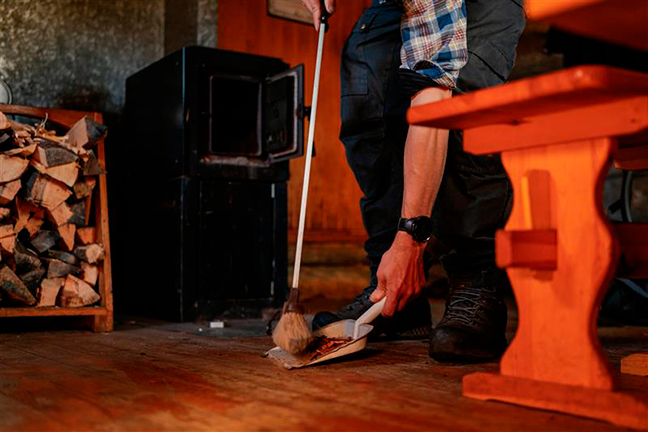 A person sweeps the floor of the hut with a brush and a dustpan, and the stove can be seen in the background,
