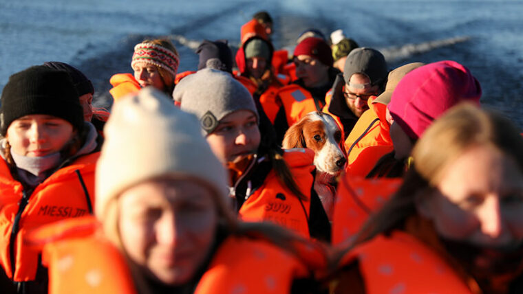 Pitk&auml;ss&auml; veneess&auml; ihmisi&auml; pelastusliivi yll&auml; sek&auml; koira. Taustalla jokimaisema.