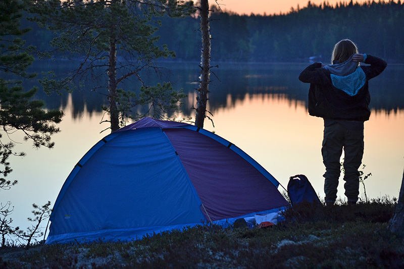 A camper stands by the shore next to their tent on a summer evening.