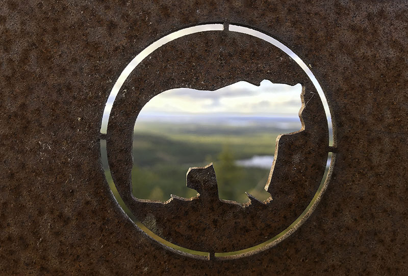 I en metallplåt har ett björnformat hål skurits ut, och genom hålet syns ett landskap med älv och skog.