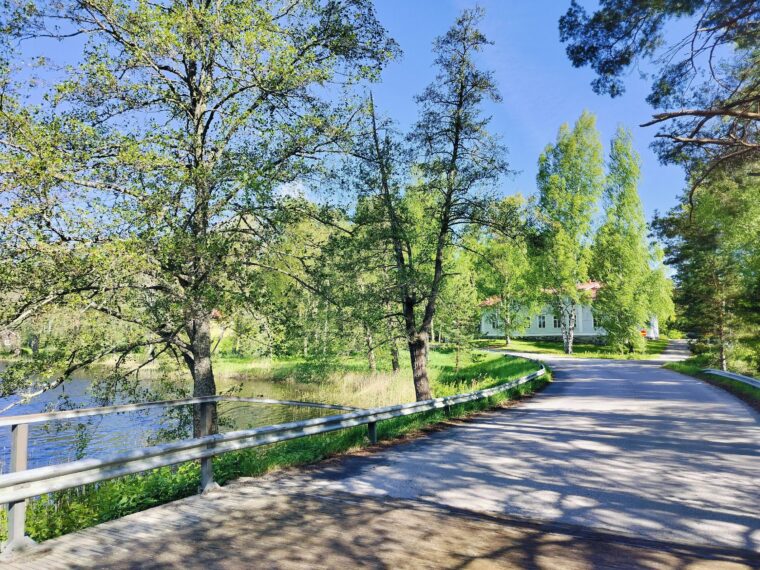 Kapea asfaltoitu tie k&auml;&auml;ntyy mutkaan. Tiell&auml; on reunakaiteet. Toisella puolella tiet&auml; n&auml;kyy vett&auml; ja mutkan takana vanha iso talo.