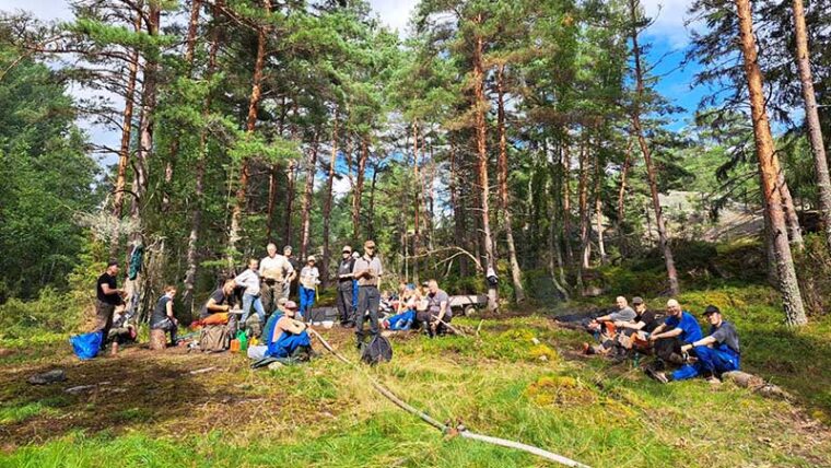 Ihmisjoukko tauolla pienellä mäntyjen reunustamalla aukolla. Ihmiset katsovat kohden kameraa. Osa istuu ja osa seisoo.