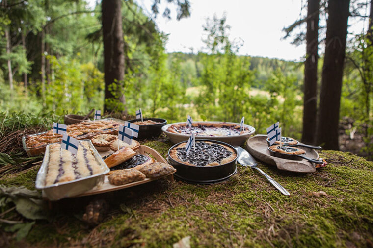 Erilaisia mustikkapiirakoita on aseteltu tarjolle kakkulapion kera sammaleiselle alustalle metsässä. Piirakat on koristeltu Suomen lipuilla.