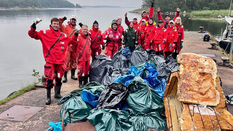 Rannalla on joukko ihmisi&auml; ja heid&auml;n edess&auml;&auml;n on kasa roskapusseja.