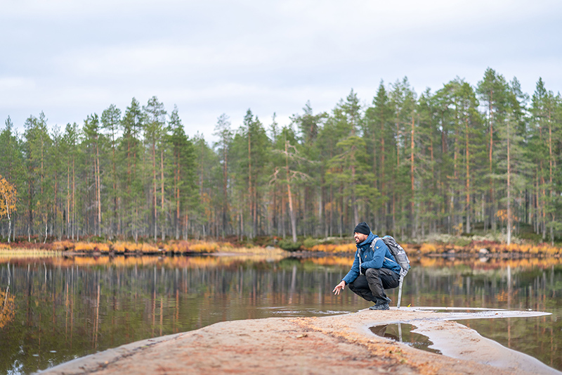 Retkeilijä on kyykyssä rannalla ja katsoo järvelle.