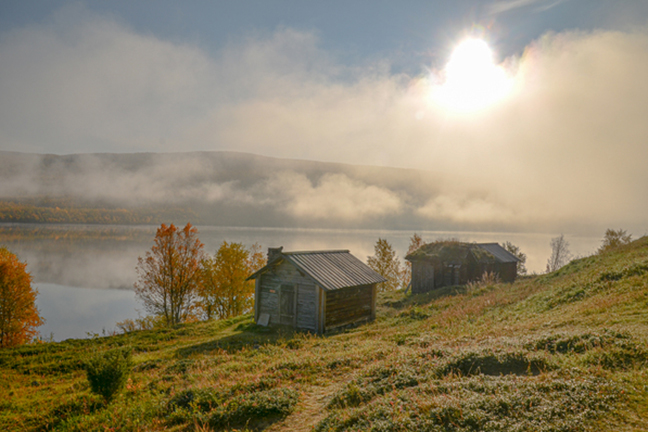 Kaksi vanhaa puista mökkiä veden äärellä. Taustalla syksyinen maisema ja usvaa.