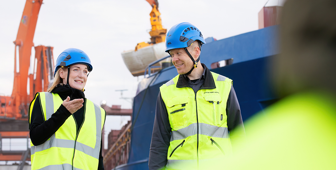 Two people are walking in the harbor wearing yellow safety vests and helmets. Cranes and ships are visible in the background.