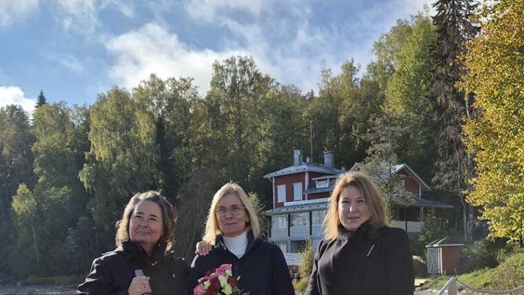 Kolme henkilöä seisoo laiturilla taustallaan kaksikerroksinen puurakennus.