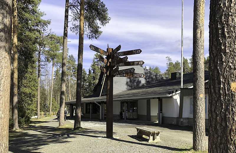 Vaalean rakennuksen sisäänkäynnin edustalla on liuskekivilaatta. Piha on hiekkaa ja siinä kasvaa suuria mäntyjä. Etualalla on pylväs, jossa on paljon opasteviittoja.