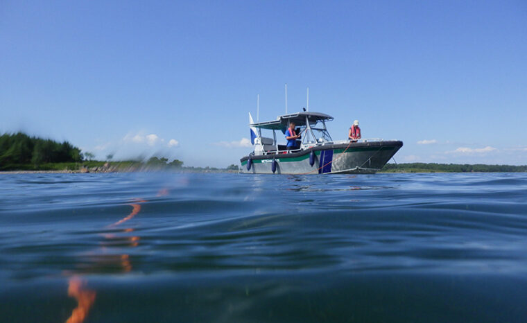Kaksi henkilöä moottoriveneessä. Kuva on otettu vedenpinnan tasolta ja kuvan etualalla on veden alla oleva köysi.